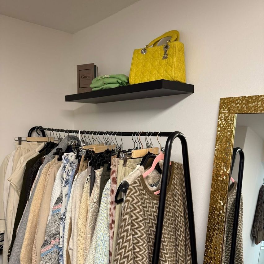 Ein Raum mit einem Kleiderständer voller verschiedener Outfits und einer gelben Handtasche auf einem Regal darüber. Ein goldener Spiegel wird auf der rechten Seite reflektiert.