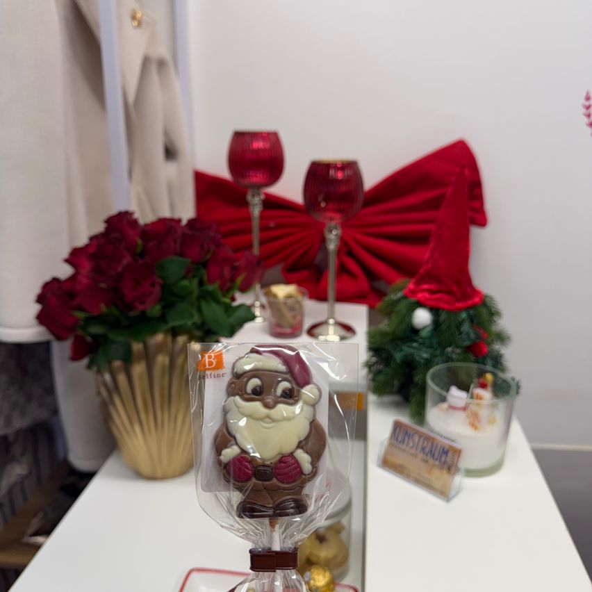 Auf einem Tisch sind eine Schokoladenfigur von Santa Claus, rote Weinkelche und ein Strauß roter Rosen zu sehen. Eine rote Schleife, ein Weihnachtsbaum und eine kleine Kerze sind ebenfalls vorhanden.