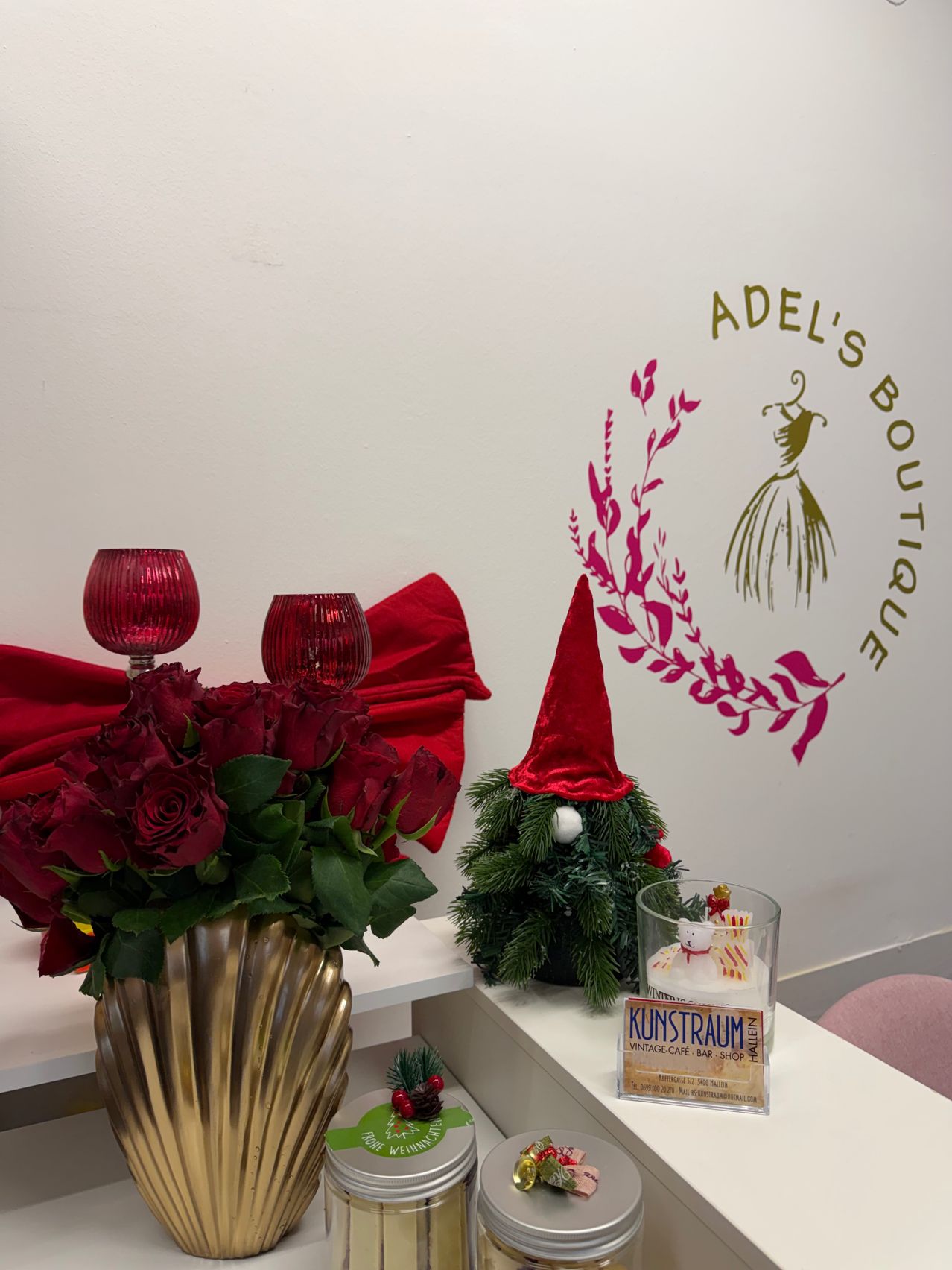 Ein weißer Regal präsentiert eine goldene Vase mit roten Rosen, einem roten Gnom und einer Kerze. Eine weiße Wand hat ein rosa Logo mit 'Adel's Boutique'. Ein Glasbecher hat einen kleinen Schneemann und andere Gegenstände.