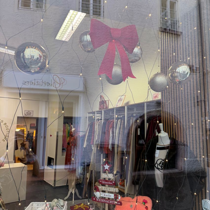 Ein Schaufenster zeigt eine weihnachtliche Dekoration mit Kugeln, einer großen roten Schleife und dekorativen Lichtern. Die Reflexion zeigt eine Puppe, ein Sternschild und Kleidungsstücke.