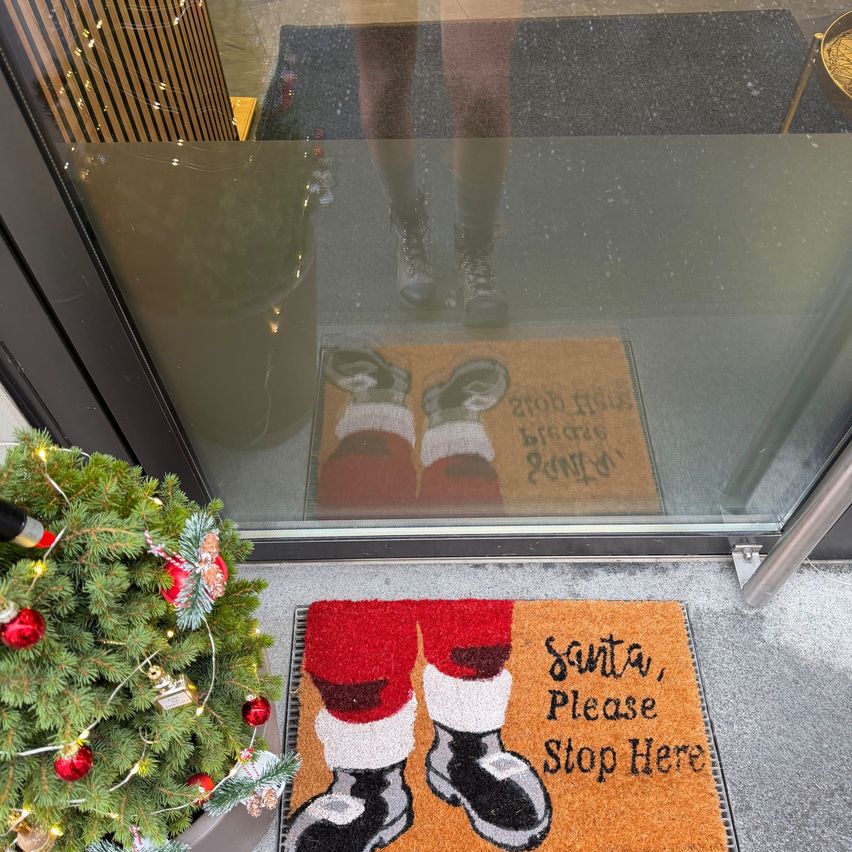 Ein festlicher Weihnachtsteppich mit Santas Stiefeln liegt auf dem Boden. Der Teppich trägt die Aufschrift 'Santa, bitte halten Sie hier an'. Die Reflexion einer Person ist in der Glastür sichtbar. Ein mit Lichtern geschmückter Weihnachtsbaum steht links.