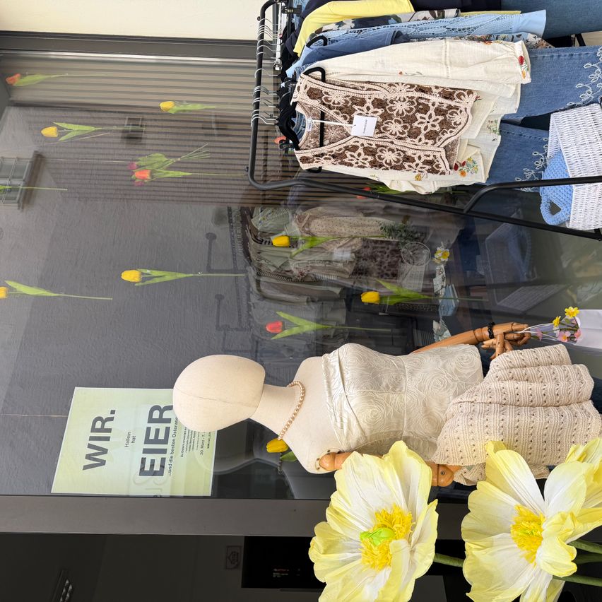 In einem Schaufenster eines Ladens sind verschiedene Kleidungsstücke an Kleiderbügeln und eine Puppe in einem weißen Kleid zu sehen, mit einem Schild, das 'WIR. EIER' und gelbe Tulpen im Hintergrund steht.