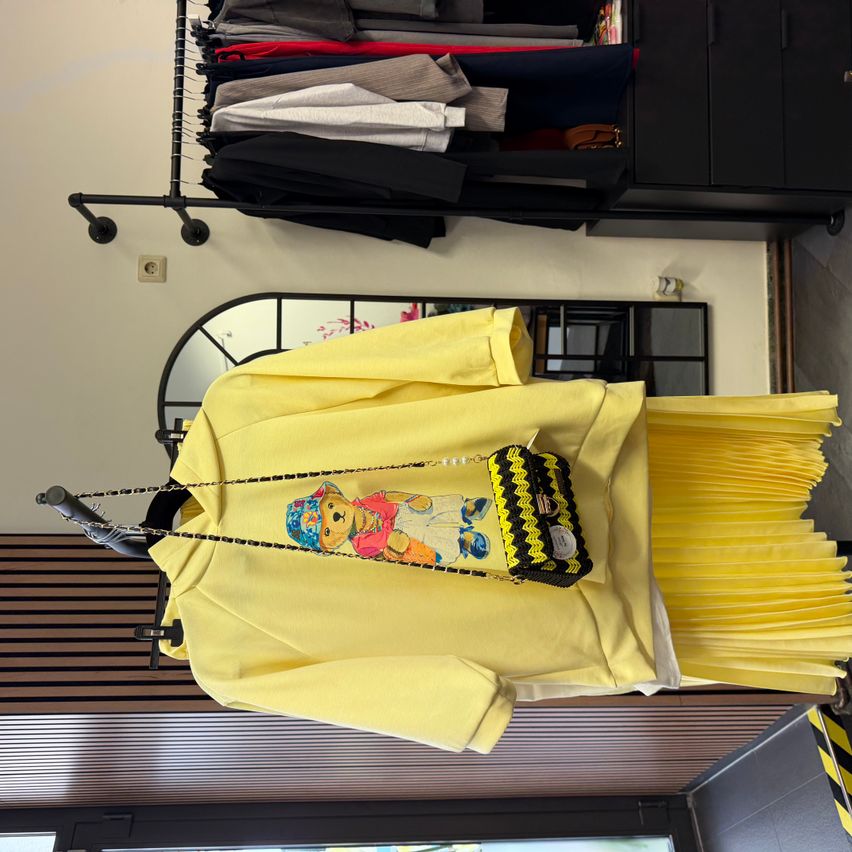 Ein leuchtend gelbes Outfit, bestehend aus einer Jacke, einem Rock und einer schwarzen Handtasche mit Bärendesign, hängt an einem Kleiderständer über einem Spiegel.