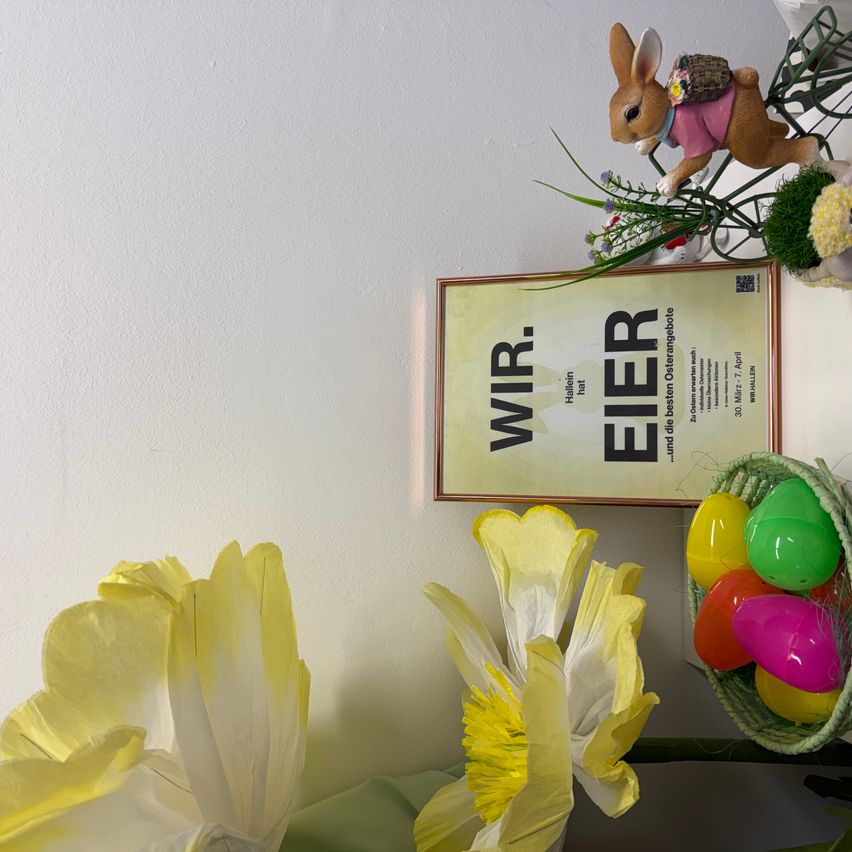 Ein Poster mit dem Titel Wir Eier ist an einer weißen Wand befestigt. Darunter befinden sich Ostereier und eine Blume in einem Korb. Es gibt auch eine Papierblume und eine Kaninchen-Spielzeugfigur mit einem Korb auf dem Rücken.