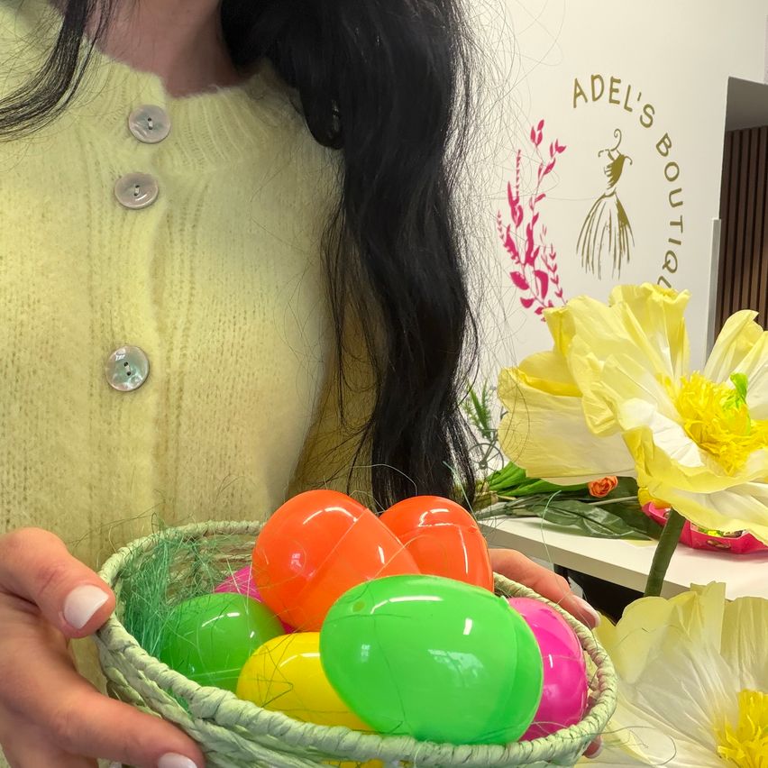 Eine Frau mit langen schwarzen Haaren hält einen Korb mit bunten Plastikeiern. Der Korb ist grün mit einem geflochtenen Design. Im Hintergrund befindet sich eine gelbe Blume und das Schild Adel's Boutique.