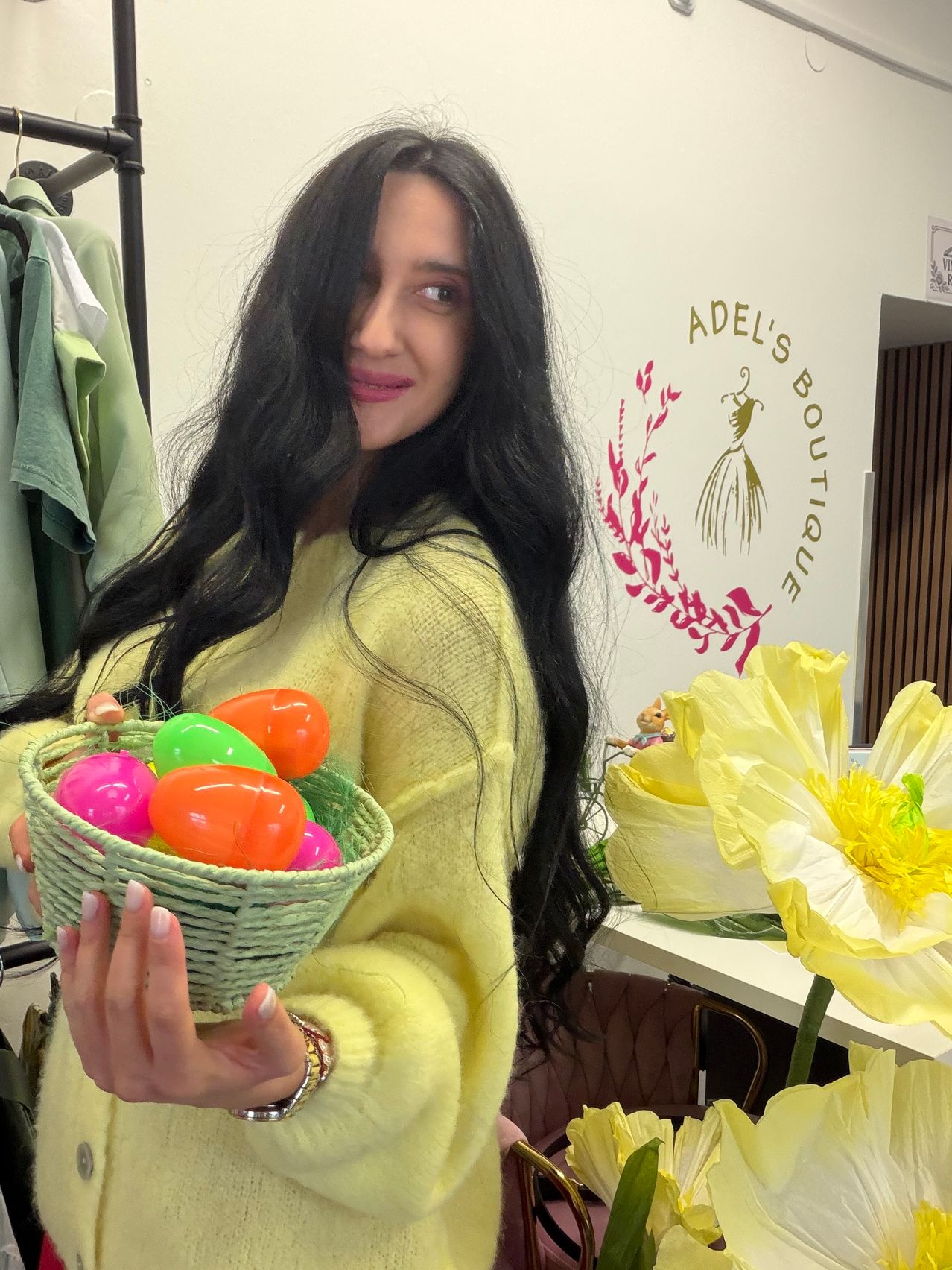 Eine Frau mit langen schwarzen Haaren hält einen Korb mit bunten Ostereiern vor einer weißen Wand mit einem Logo.