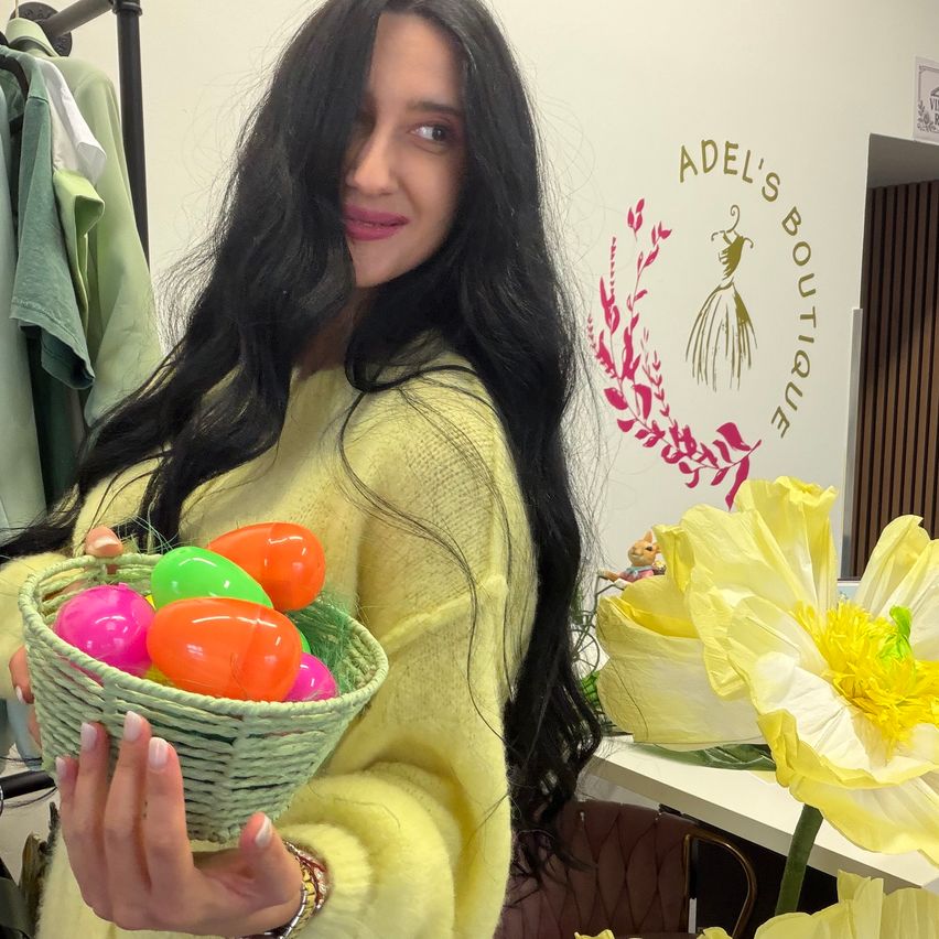 Eine Frau mit langen schwarzen Haaren hält einen Korb mit bunten Ostereiern vor einer weißen Wand mit einem Logo.