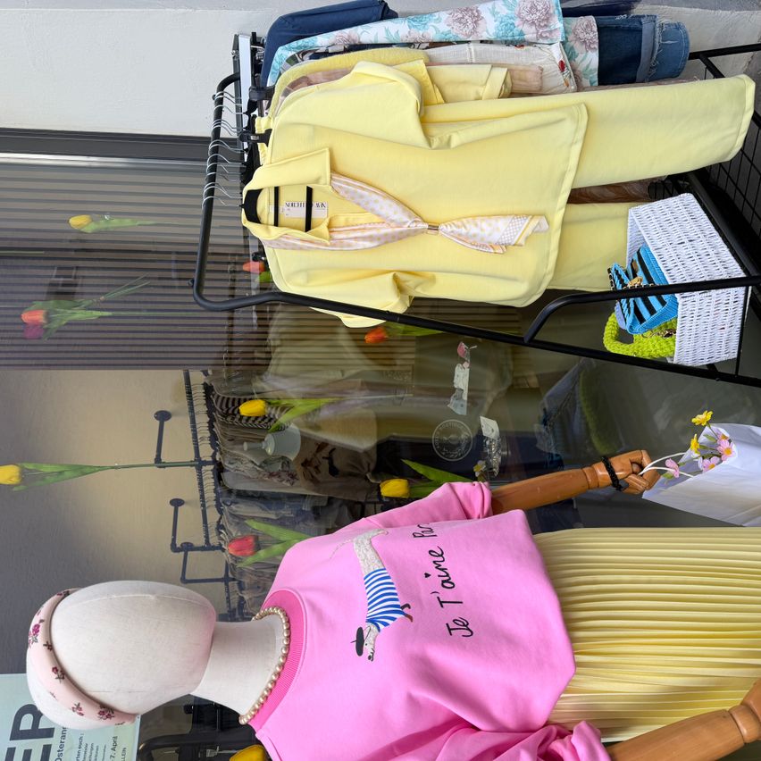 Ein Mannequin in einem Boutiquefenster zeigt ein pinkes T-Shirt mit einem französischen Satz und einem gelben Outfit. Ein Korb und andere Kleidungsstücke befinden sich auf einem Gestell dahinter.