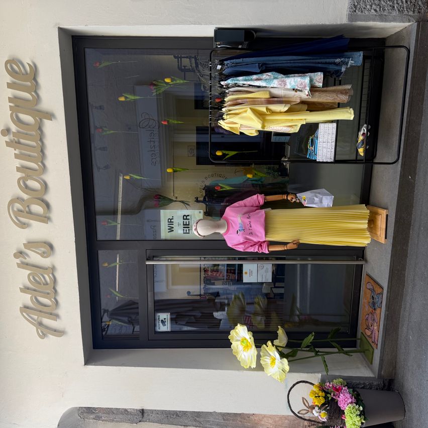 Ein Boutique-Laden zeigt eine Mannequin, die ein rosa Hemd und einen gelben Rock trägt, vor einem Fenster. Der Laden hat ein Schild mit der Aufschrift Adel's Boutique und ist mit Blumen dekoriert.