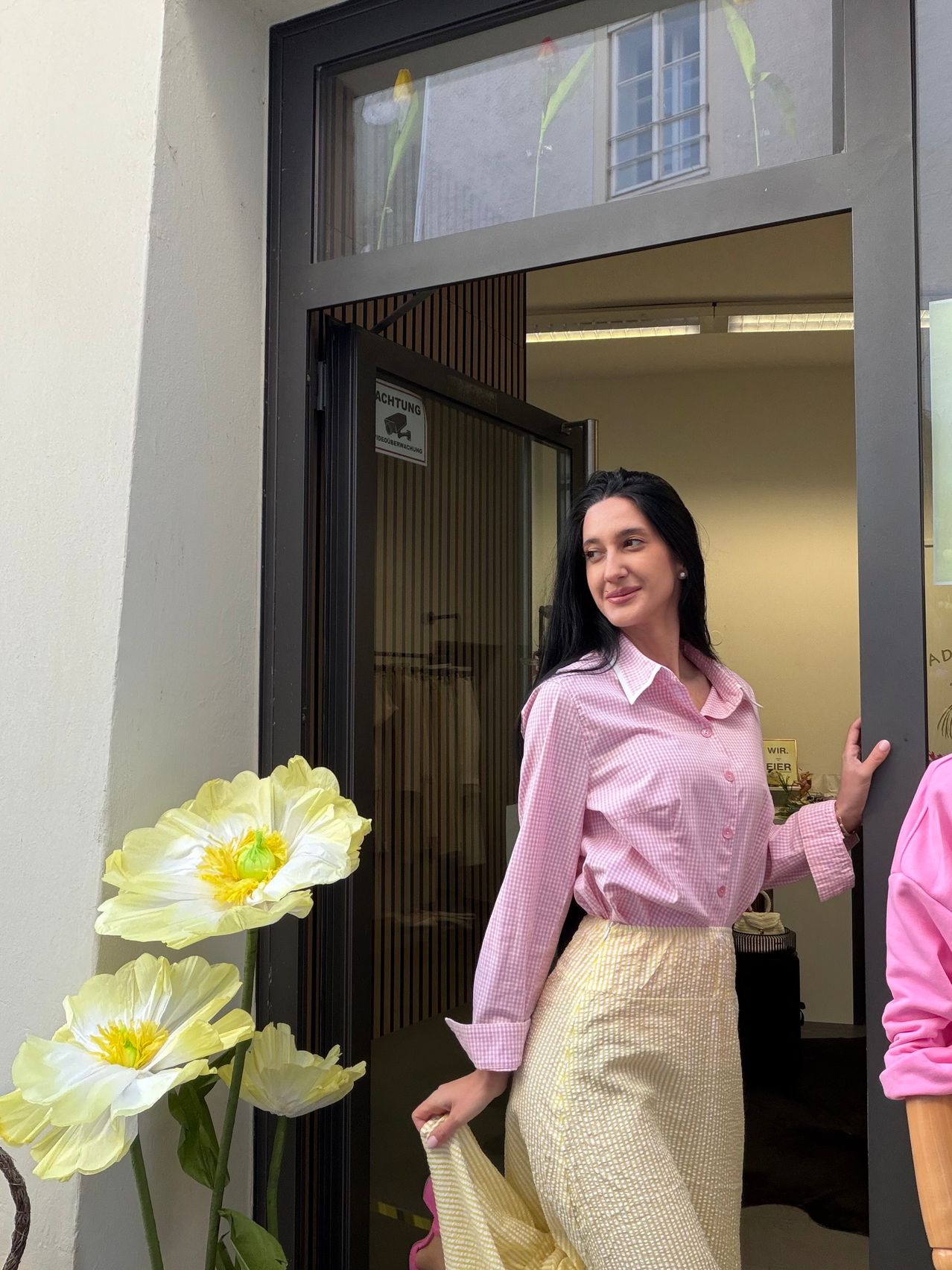 Eine Frau in einem rosa karierten Hemd und gelbem Rock posiert vor einem Laden mit Überwachungsschild, Blumen und einer offenen Tür, die in einen beleuchteten Innenraum führt.