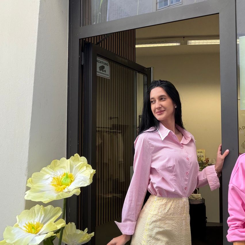 Eine Frau in einem rosa karierten Hemd und gelbem Rock posiert vor einem Laden mit Überwachungsschild, Blumen und einer offenen Tür, die in einen beleuchteten Innenraum führt.