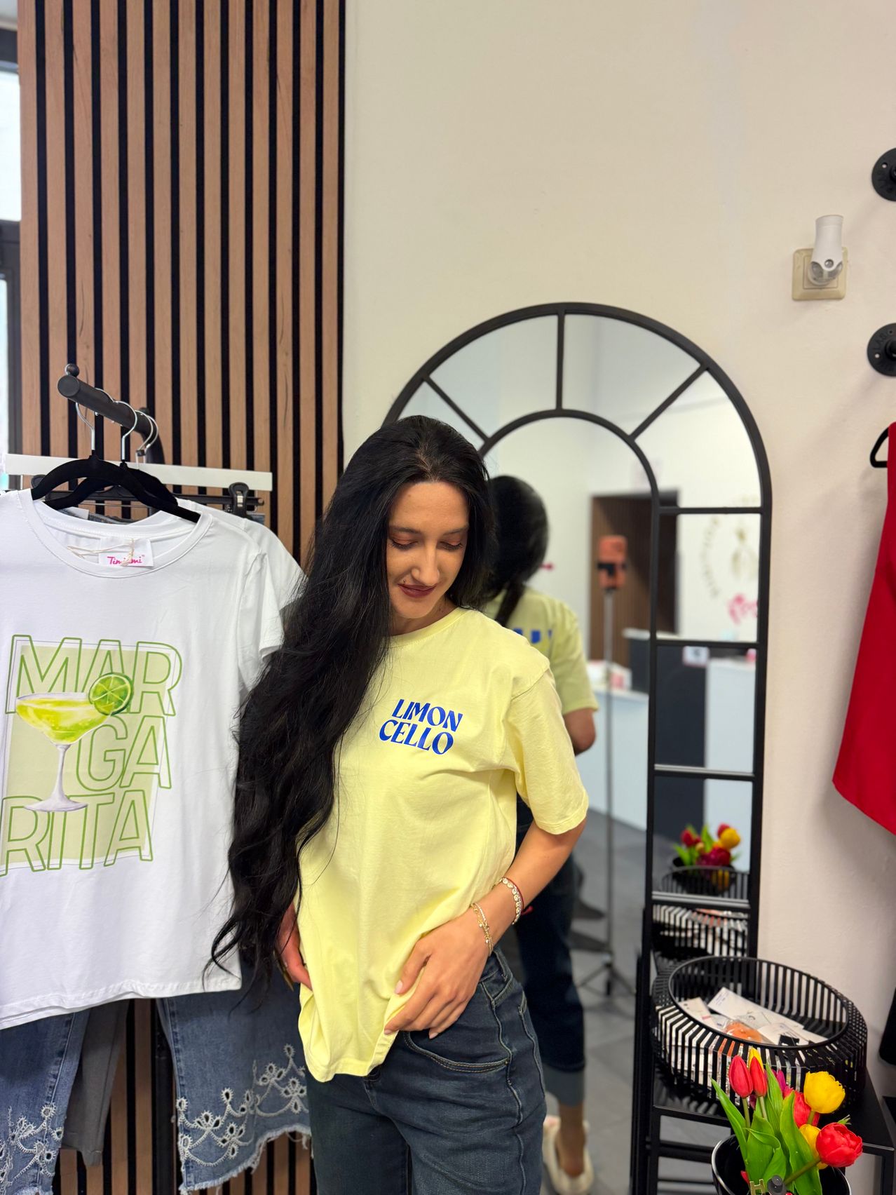 Eine Frau mit langen Haaren steht in einem Raum und lächelt. Sie trägt ein gelbes T-Shirt mit der Aufschrift Limon Cello. Ein weißes T-Shirt mit einem Cocktaildesign hängt hinter ihr. Im Spiegel ist die Reflexion einer anderen Person zu sehen.