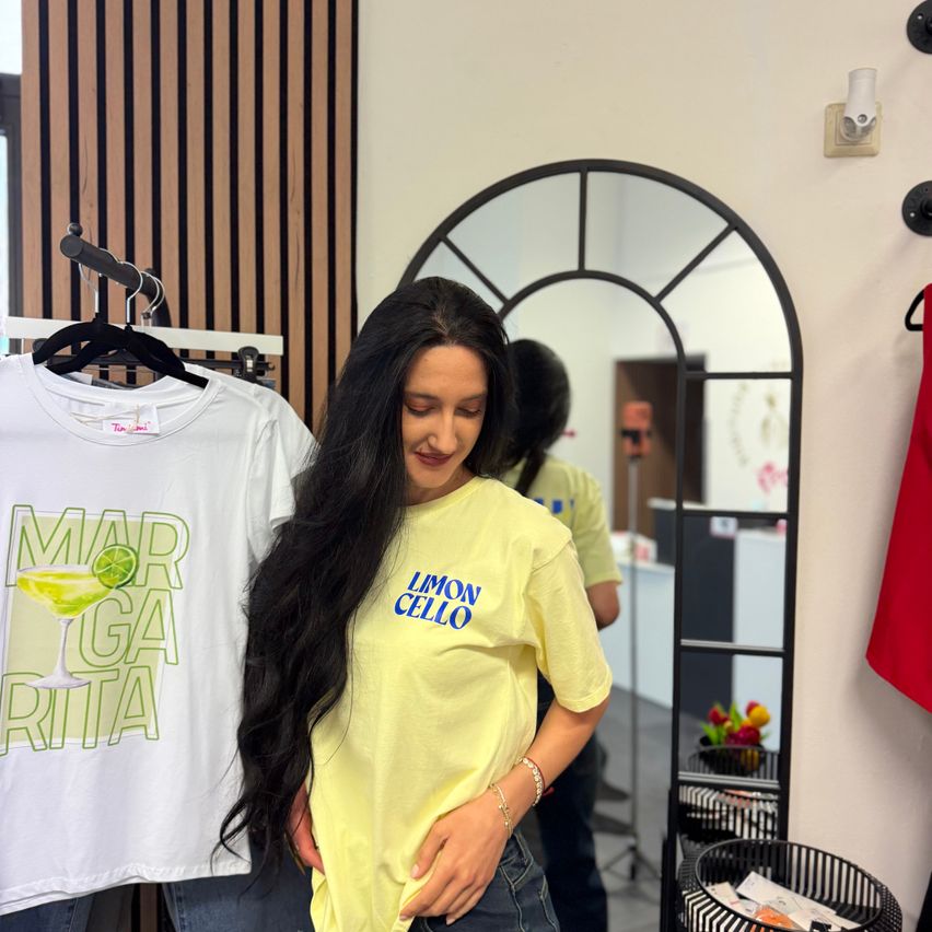 Eine Frau mit langen Haaren steht in einem Raum und lächelt. Sie trägt ein gelbes T-Shirt mit der Aufschrift Limon Cello. Ein weißes T-Shirt mit einem Cocktaildesign hängt hinter ihr. Im Spiegel ist die Reflexion einer anderen Person zu sehen.