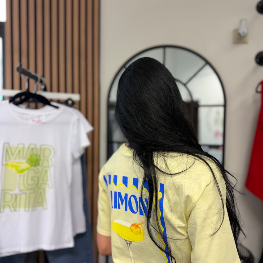 Eine Frau mit langen schwarzen Haaren schaut ein gelbes T-Shirt mit der Aufschrift 'Margarita' in einem Bekleidungsgeschäft an.