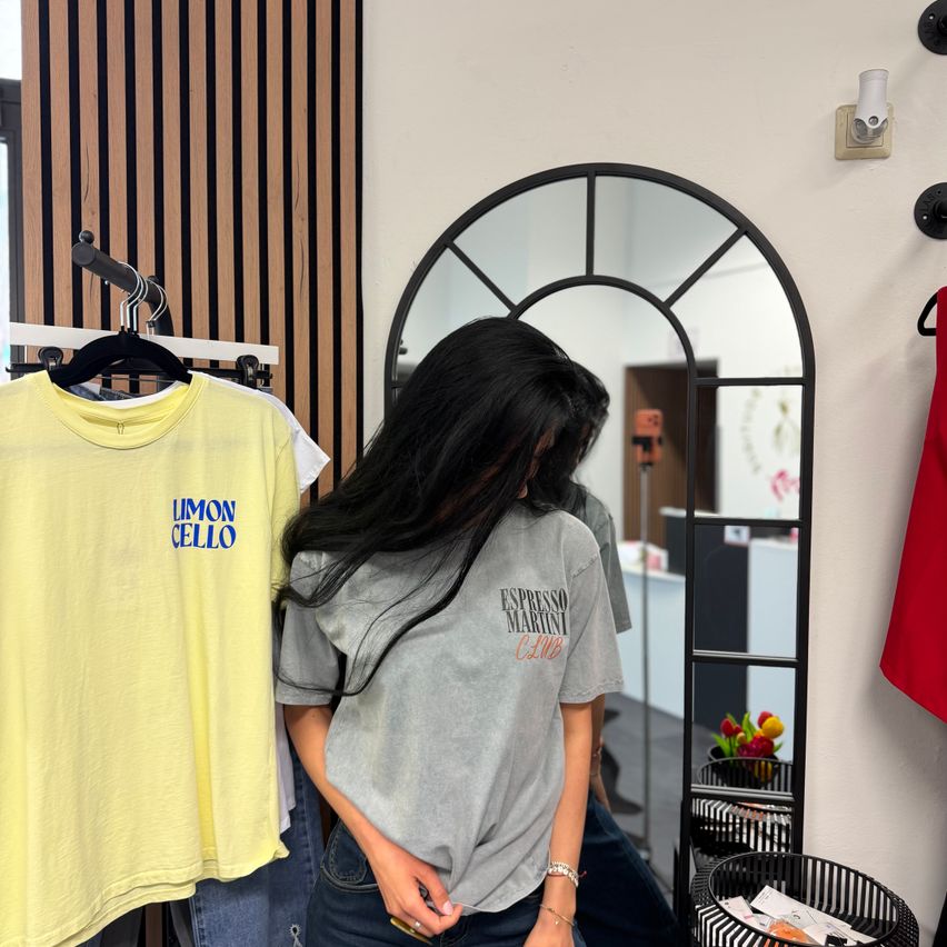 Eine Frau betrachtet ein gelbes T-Shirt mit der Aufschrift 'LIMON CELLO' vor einem Spiegel in einem Bekleidungsgeschäft. Sie trägt ein graues Shirt mit der Aufschrift 'ESPRESSO MARINO CAFE'.
