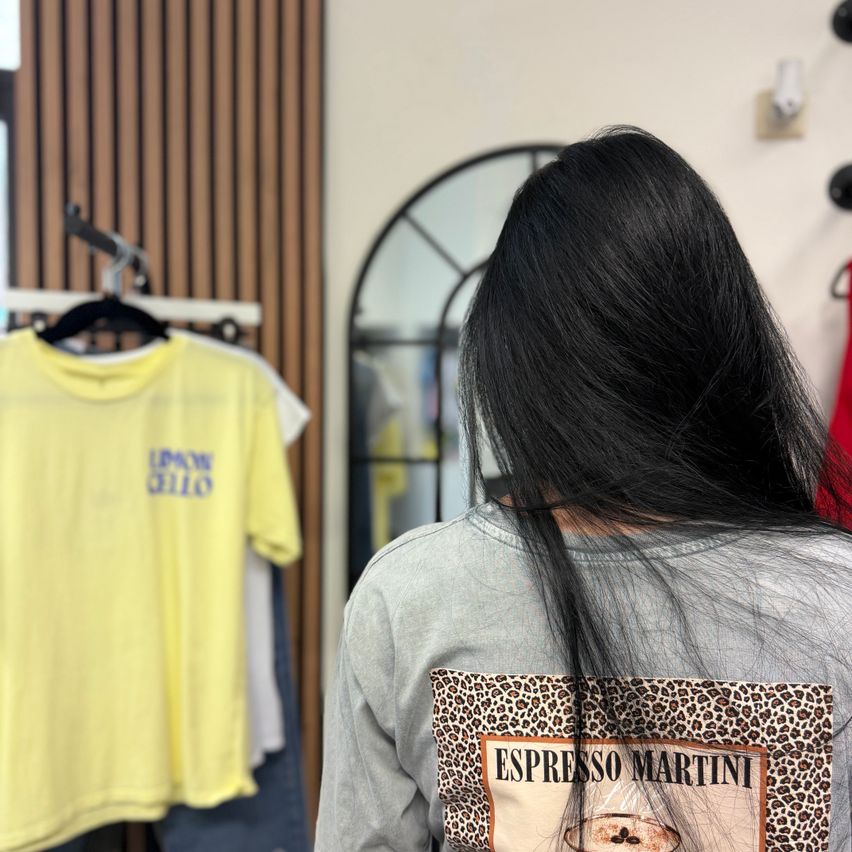 Eine Frau mit langen Haaren steht in einem Bekleidungsgeschäft. Vor ihr ist ein graues T-Shirt mit einem Espresso Martini-Grafik und hinter ihr ist ein gelbes T-Shirt an einem Kleiderbügel.