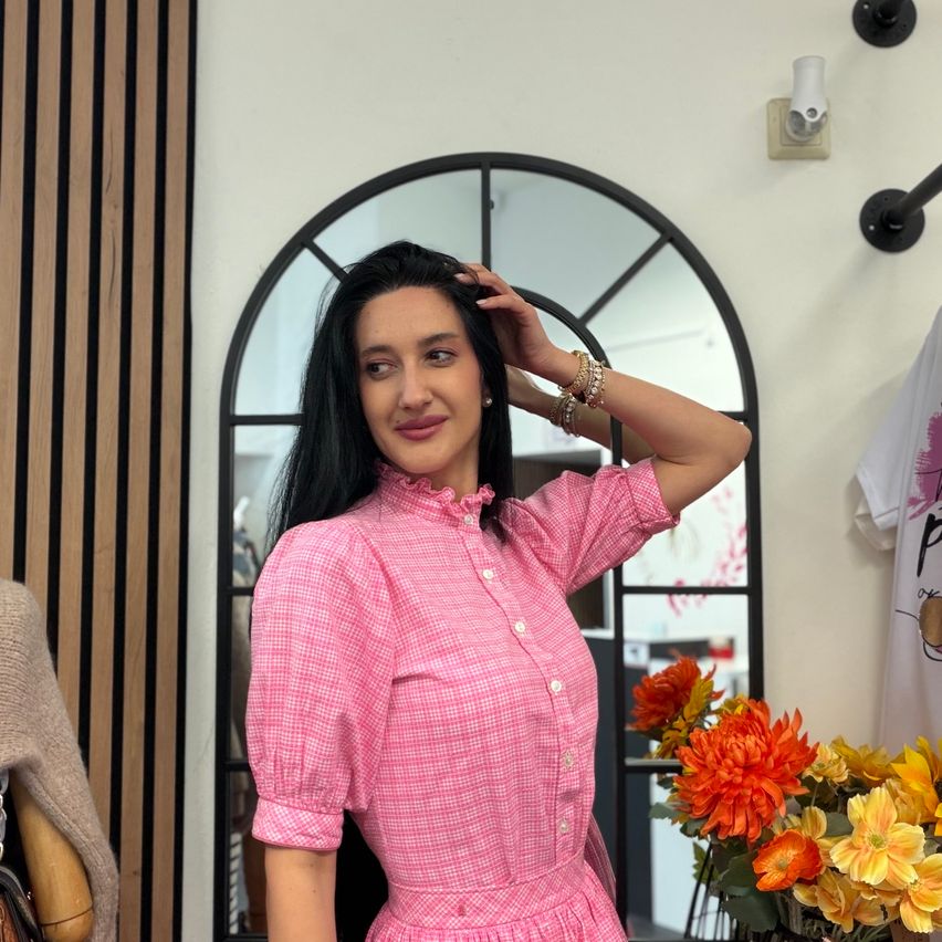 Eine Frau steht vor einem Spiegel und trägt ein rosa kariertes Kleid. Sie berührt ihr Haar und lächelt. Dahinter ist eine Holzplatte, ein weißes Hemd und ein Blumenstrauß.