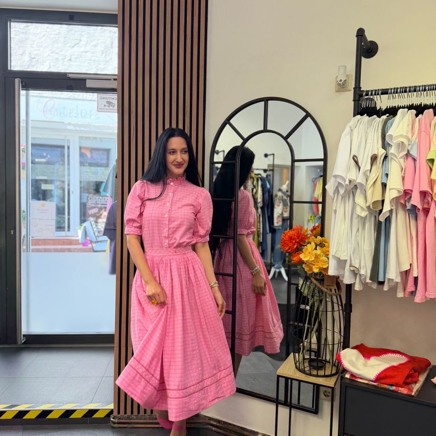 Eine Frau in einem rosa Kleid steht vor einem Spiegel in einem Bekleidungsgeschäft. Bunte Kleidung hängt auf einem Kleiderständer in der Nähe.