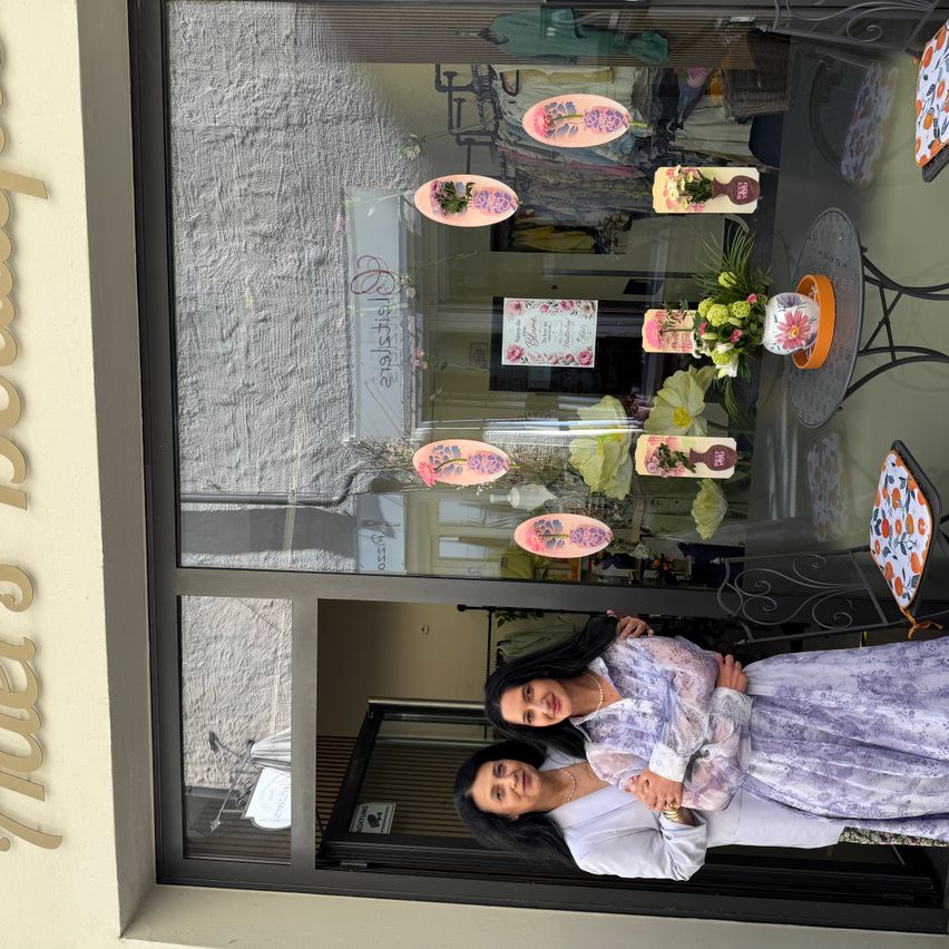 Zwei Frauen stehen lächelnd und posieren vor einem Schaufenster. Der Laden hat Blumendekorationen und eine offene Tür.
