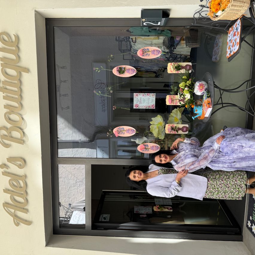 Zwei Frauen posieren vor einem Schaufenster mit Blumendekorationen. Das Schild des Ladens lautet Adele's Boutique.