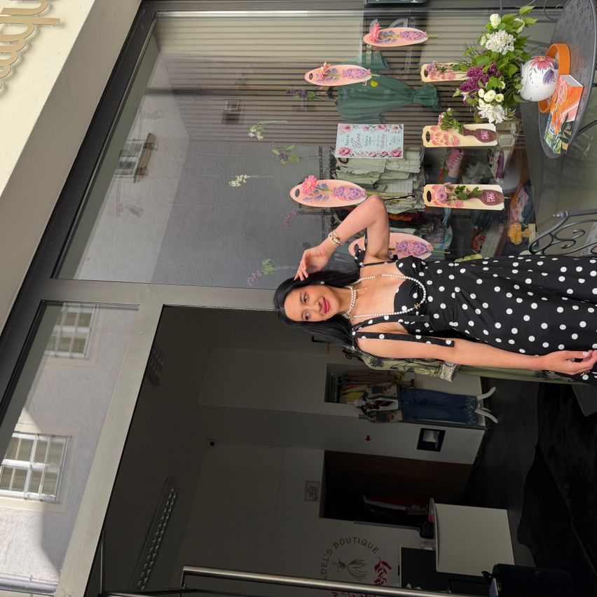 Eine Frau in einem gepunkteten Kleid steht in einem Geschäft und posiert für ein Foto. Das Geschäft hat Glaswände und zeigt verschiedene Artikel. Blumen und Dekorationsgegenstände sind im Inneren zu sehen.