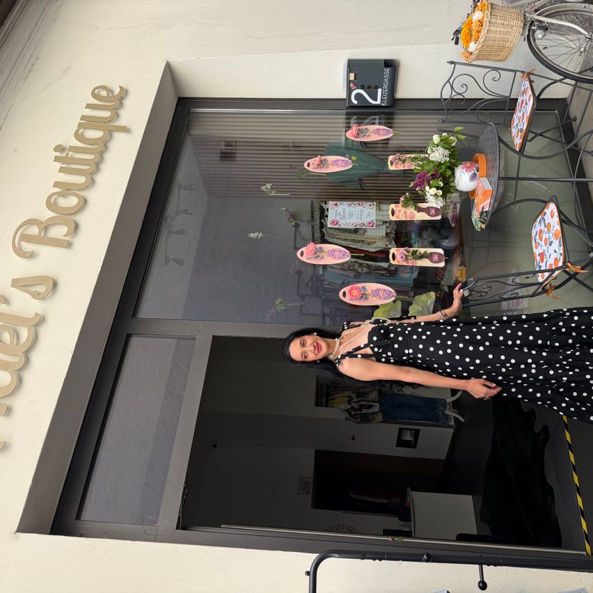Eine Frau in einem gepunkteten Kleid steht vor einem Geschäft namens A Boutique. Im Schaufenster des Geschäfts sind Blumen und Dekorationsartikel zu sehen.