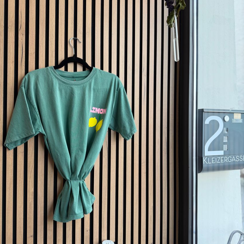 Ein grünes T-Shirt mit dem Wort Limon in Pink und zwei Zitronen in Gelb hängt an einer Holzwand mit vertikalen Streifen.