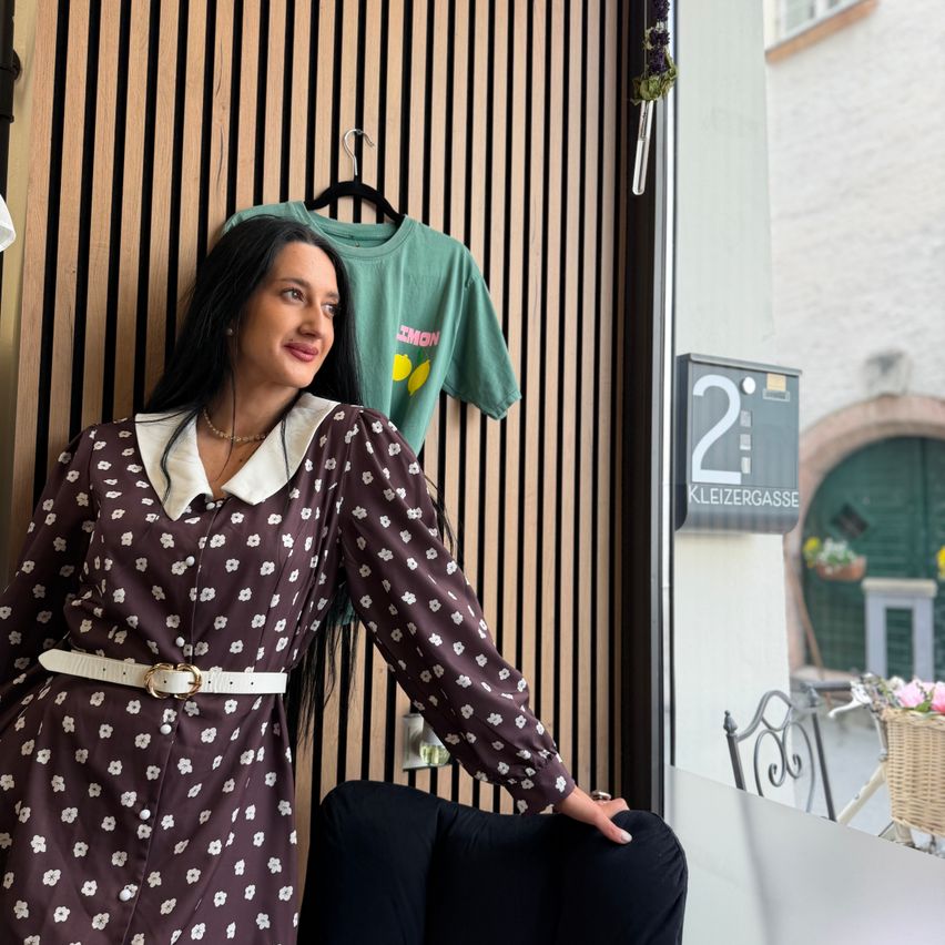 Eine Frau in einem Blumenkleid steht vor einer Holzwand mit einem grünen T-Shirt, das hinter ihr hängt. Sie lächelt und schaut in die Kamera. Das Schild mit der Nummer 2 und ein Korb sind im Fenster zu sehen.