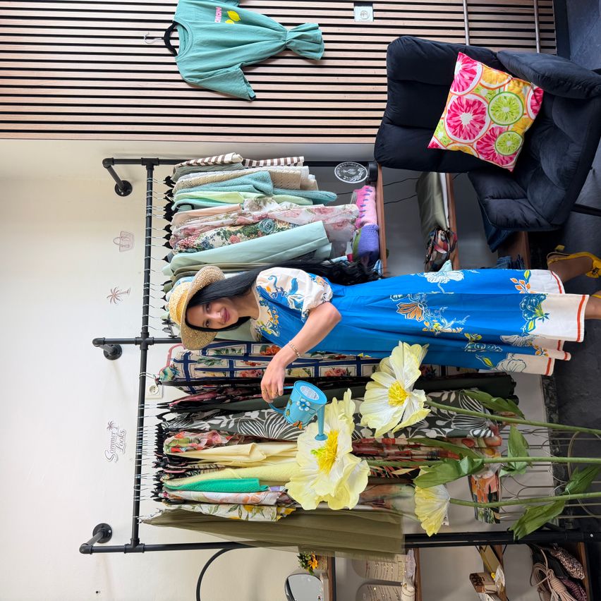 Eine Frau in einem blauen Blumenkleid und Hut gießt künstliche Blumen in einer Boutique mit einem Kleiderständer hinter ihr.