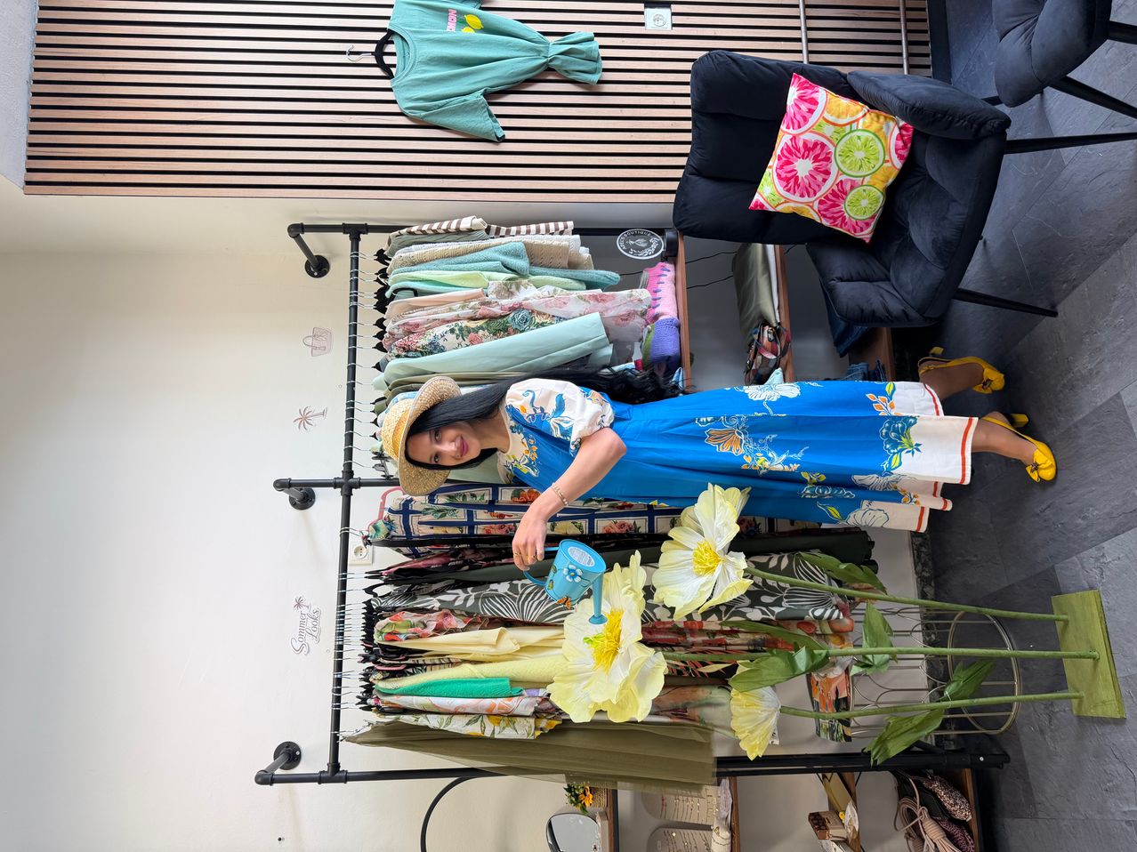 Eine Frau in einem blauen Blumenkleid und Hut gießt künstliche Blumen in einer Boutique mit einem Kleiderständer hinter ihr.