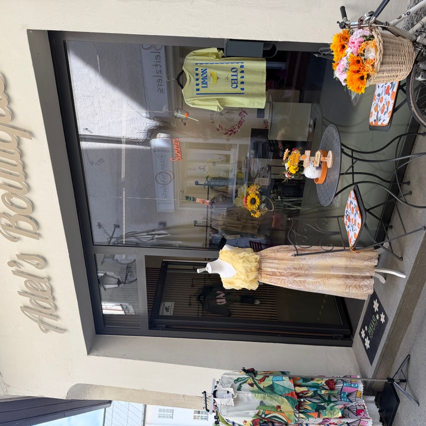 Adels Boutique-Schaufenster mit einer Glasscheibe, einem gelben Mannequin in einem Kleid, einem Tisch mit Blumen und einem Fahrrad davor.