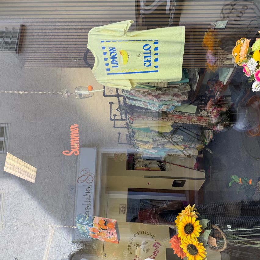 Ein Schaufenster zeigt ein gelbes T-Shirt mit Zitronen- und Cello-Aufdruck, neben einem Strauß Sonnenblumen. Eine Reflexion zeigt das Innere mit verschiedenen Artikeln.