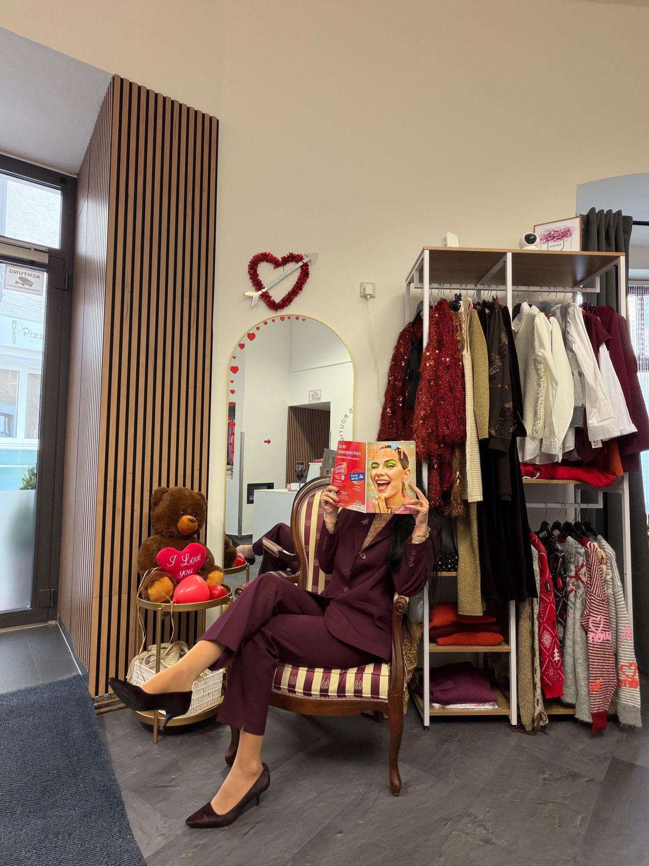 Eine Frau sitzt auf einem Stuhl und hält eine Zeitschrift in der Hand, umgeben von einem Raum mit Kleiderständern, einem Spiegel und einer herzförmigen Dekoration. Ein Teddybär mit einem Herz auf der Brust steht auf einem Tisch.