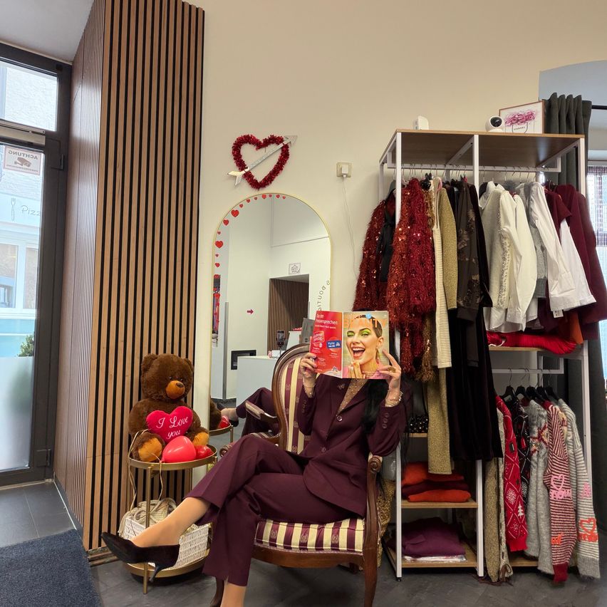 Eine Frau sitzt auf einem Stuhl und hält eine Zeitschrift in der Hand, umgeben von einem Raum mit Kleiderständern, einem Spiegel und einer herzförmigen Dekoration. Ein Teddybär mit einem Herz auf der Brust steht auf einem Tisch.