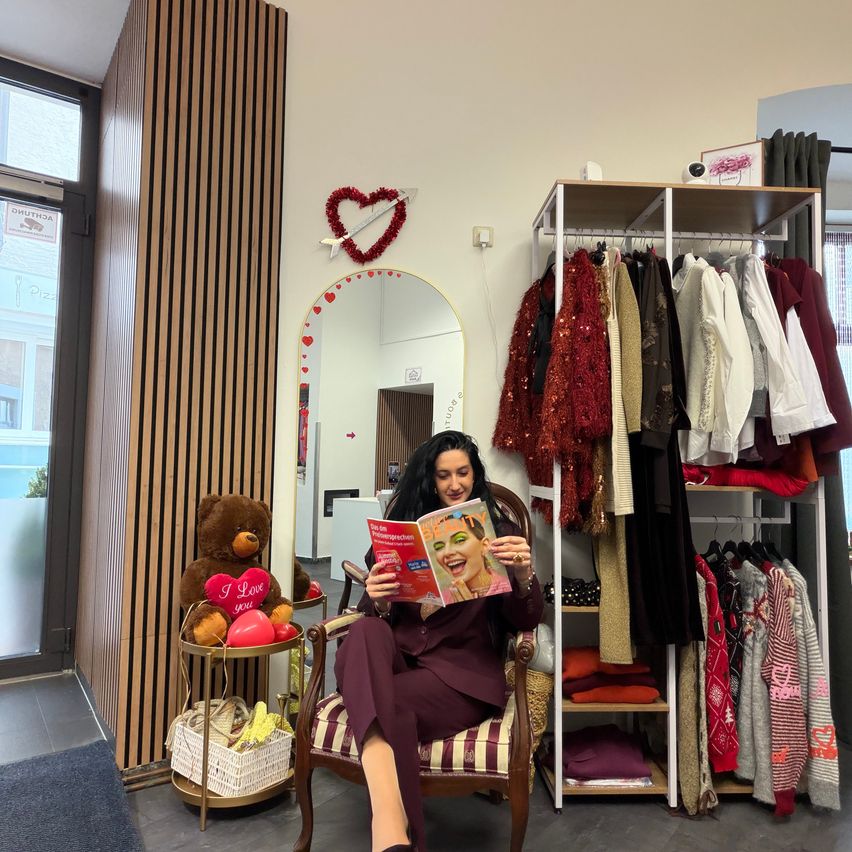 Eine Frau sitzt auf einem Stuhl, liest eine Zeitschrift und lächelt. Ein Teddybär mit einem Herz und Ballons ist neben ihr. An der Wand befindet sich ein Spiegel mit einem herzförmigen Design, und Kleidung hängt an einem Kleiderständer. Links steht ein Holzständer mit einem Korb und einem Regal.