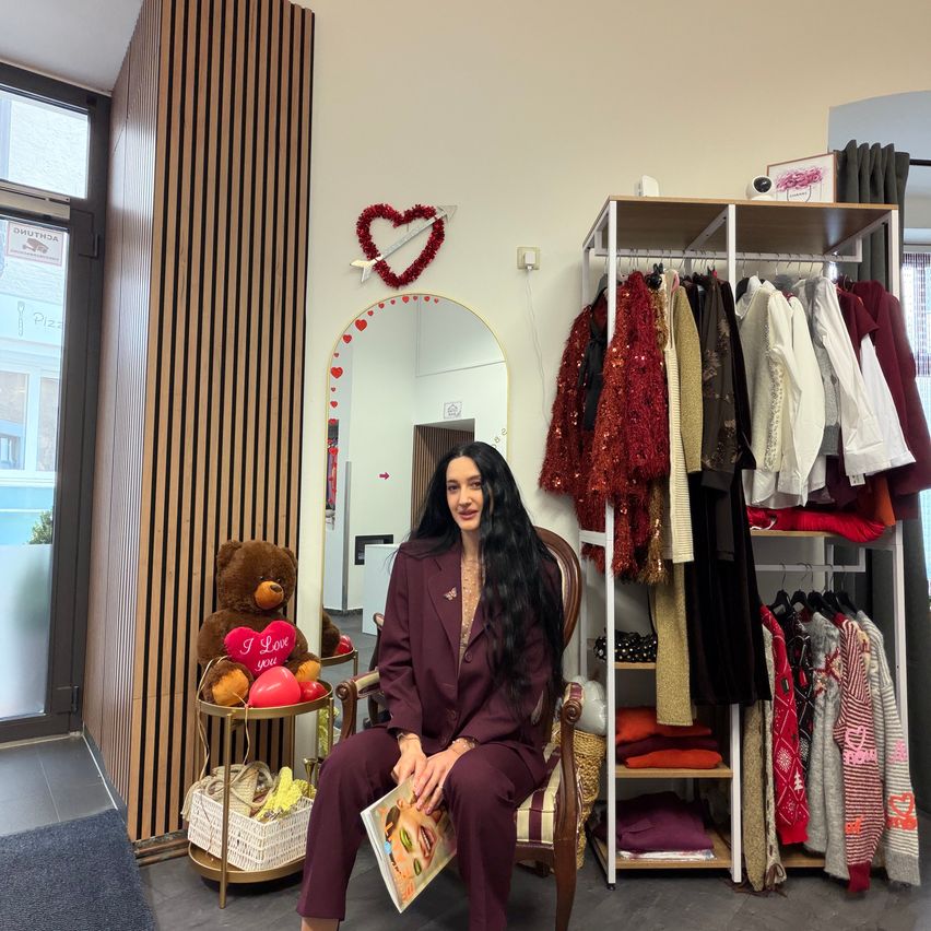 Eine Frau mit langen Haaren sitzt vor einem Kleiderständer auf einem Stuhl und lächelt. Sie hält eine Zeitschrift in der Hand und ist umgeben von einem Spiegel, einer herzförmigen Dekoration und einem Teddybären mit Luftballons.