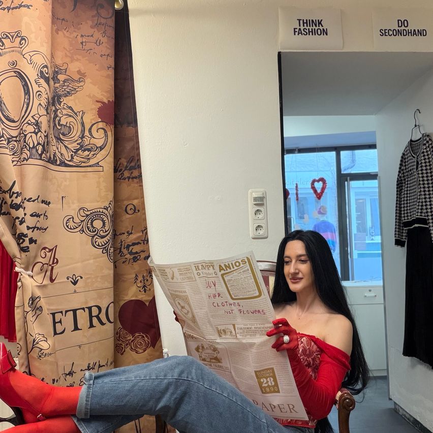 Eine Frau sitzt auf einem Stuhl und liest eine Zeitung, trägt rote Stiefel und Handschuhe. Hinter ihr befindet sich ein Vorhang mit Text und Designs. Rechts ist ein Glasfenster und ein hängendes Tuch. Oben befinden sich Schilder mit den Aufschriften 'DENKE FASHION' und 'TAGE SECONDHAND'.