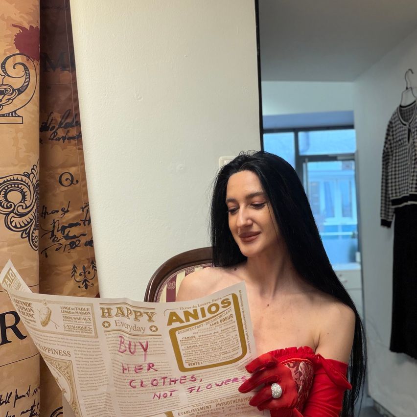 Eine Frau liest eine Zeitung mit der Aufschrift 'Happy AniOs', während sie rote Handschuhe und einen Ring trägt. Sie hat langes schwarzes Haar und steht in einem Raum mit einer weißen Wand und einem Fenster.