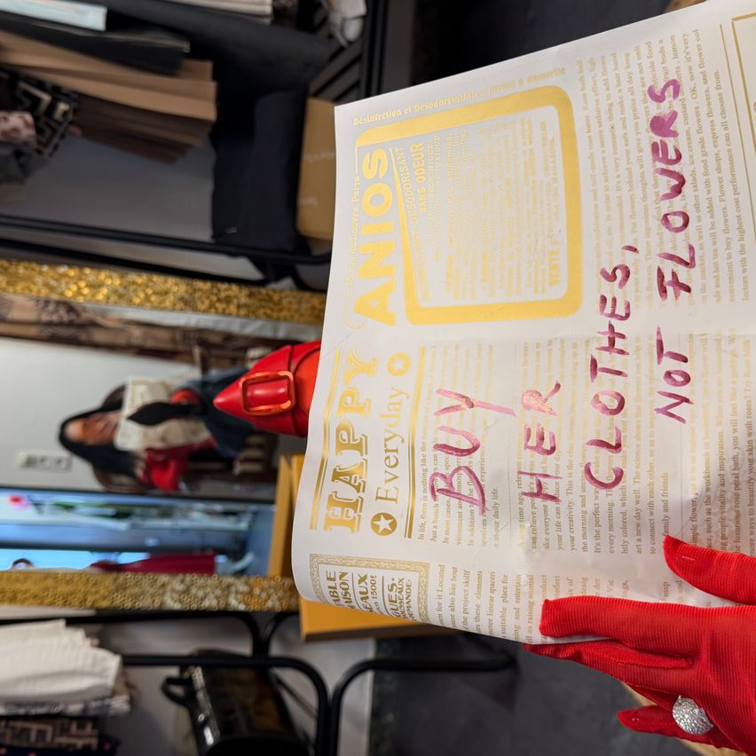 Ein Papier mit 'Happy Anos' darauf, in Gelb geschrieben. Eine Person hält es und das Papier ist gefaltet. Es scheint ein Flyer für eine Veranstaltung zu sein. Die Person trägt rote Handschuhe. Im Hintergrund befindet sich ein Spiegel und einige Objekte.