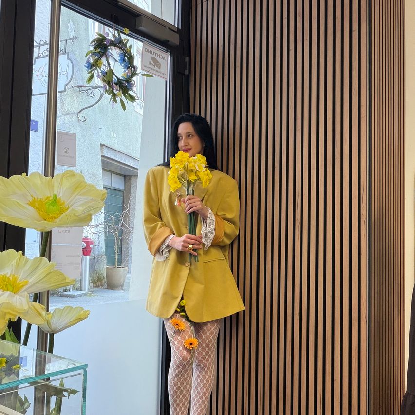 Eine Frau in einem gelben Mantel und Blumenstrümpfen steht neben einer Tür und hält einen Strauß gelber Blumen. Über der Tür hängt ein Kranz, und ein Fenster hinter ihr zeigt eine Straße mit einem Topfpflanzen.