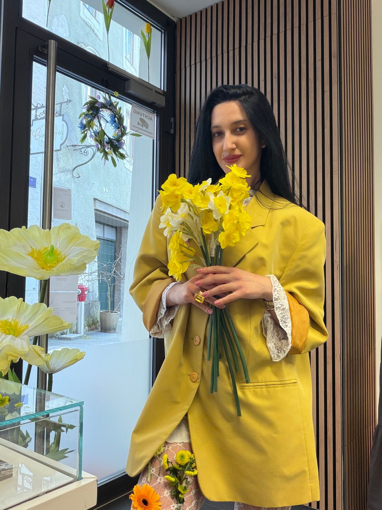 Eine Frau in einem gelben Mantel steht drinnen und hält einen Strauß gelber Narzissen. Sie lächelt, während sie in der Nähe einer Glastür steht, mit Blumen und Grünpflanzen draußen.