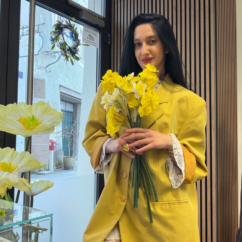 Eine Frau in einem gelben Mantel steht drinnen und hält einen Strauß gelber Narzissen. Sie lächelt, während sie in der Nähe einer Glastür steht, mit Blumen und Grünpflanzen draußen.