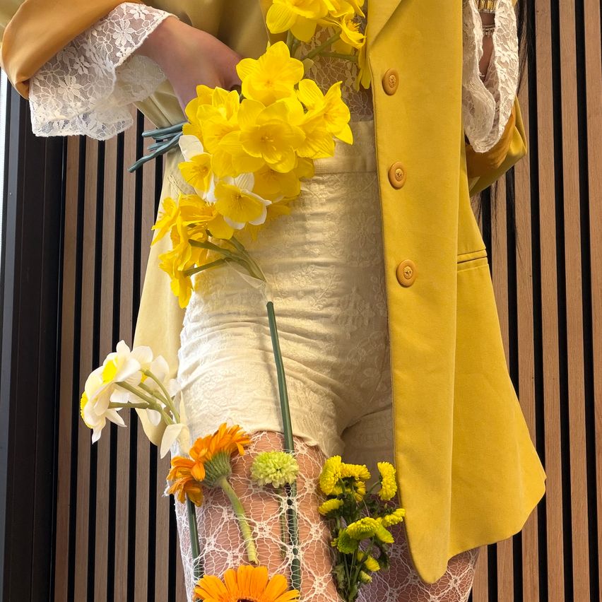 Eine Frau in einem gelben Mantel mit Spitzenbesatz hält einen Strauß gelber und weißer Blumen und trägt passende Spitzenshorts und Strümpfe.