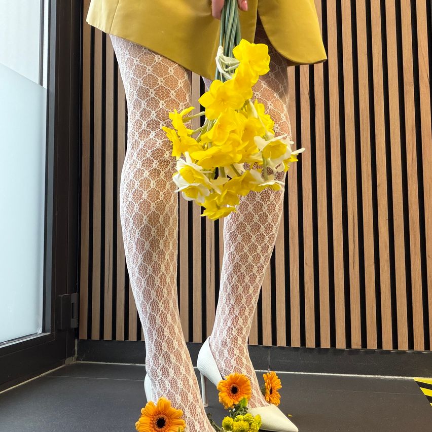 Eine Person steht mit einem Strauß gelber Blumen vor einer Holzwand, trägt gemusterte Spitzenstrümpfe und gelbe Schuhe.