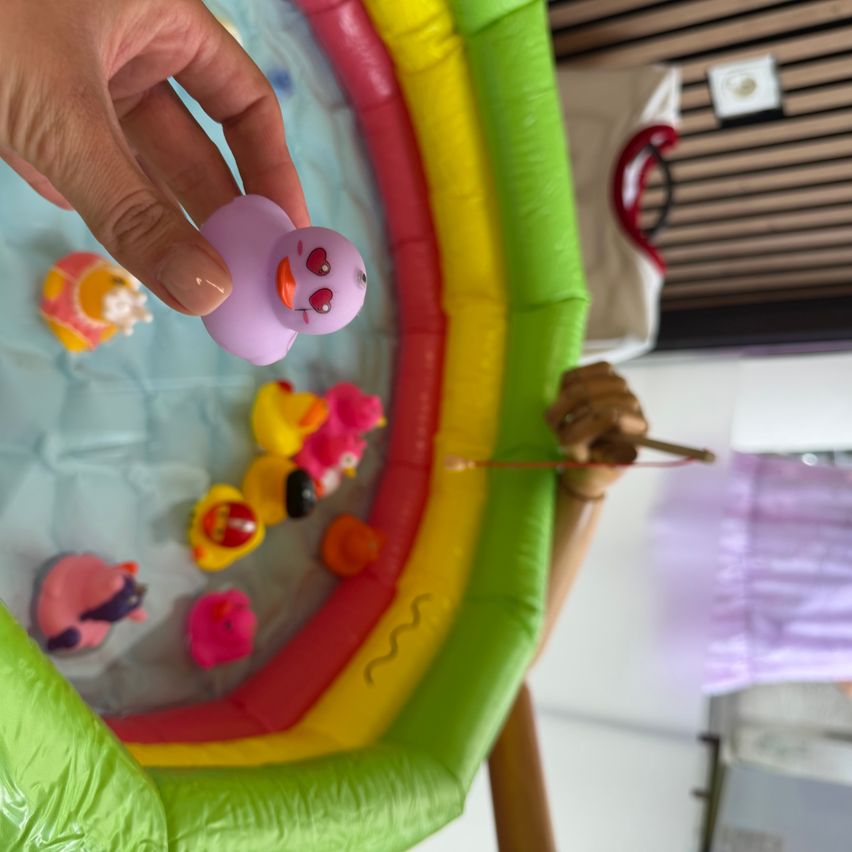 Eine Hand hält eine lila Gummiente in einem bunten aufblasbaren Pool mit anderen Spielzeugen.