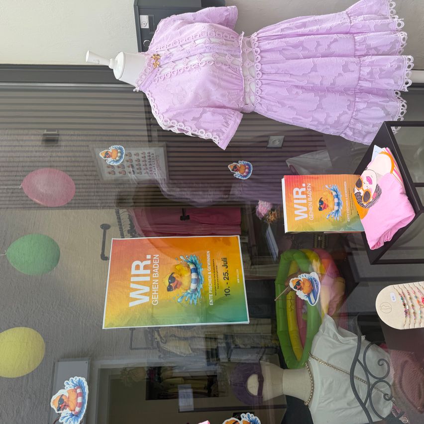 Ein Vitrinendisplay mit einem lila Kleid, einem Buch und verschiedenen bunten Gegenständen, darunter Luftballons und Aufkleber.