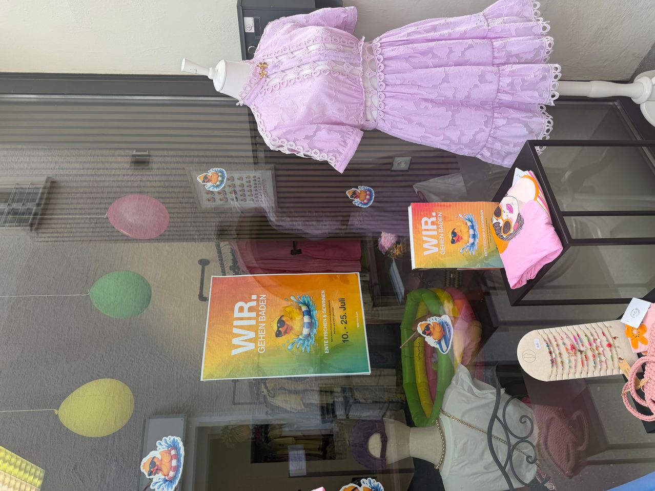 Ein Vitrinendisplay mit einem lila Kleid, einem Buch und verschiedenen bunten Gegenständen, darunter Luftballons und Aufkleber.