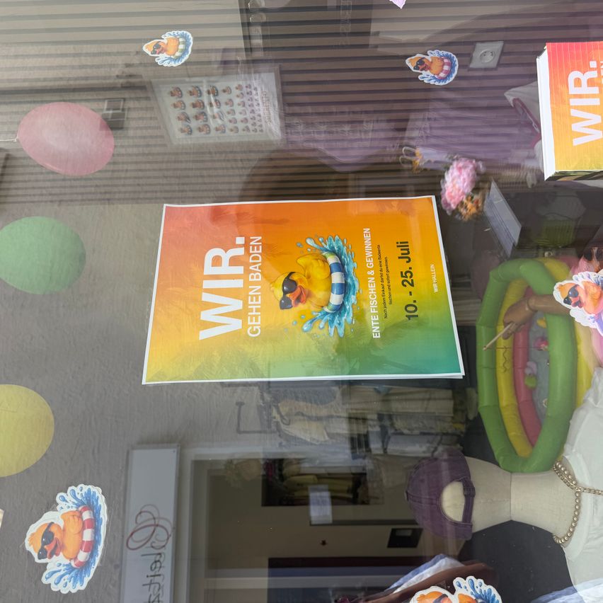 Ein Poster mit dem Text 'WIR GEHEN BADEN' und einer Comic-Figur wird auf einer Glasscheibe ausgestellt. Das Poster ist von verschiedenen Aufklebern und Luftballons umgeben. Das Datum '10-25. Juli' ist ebenfalls auf dem Poster angegeben.