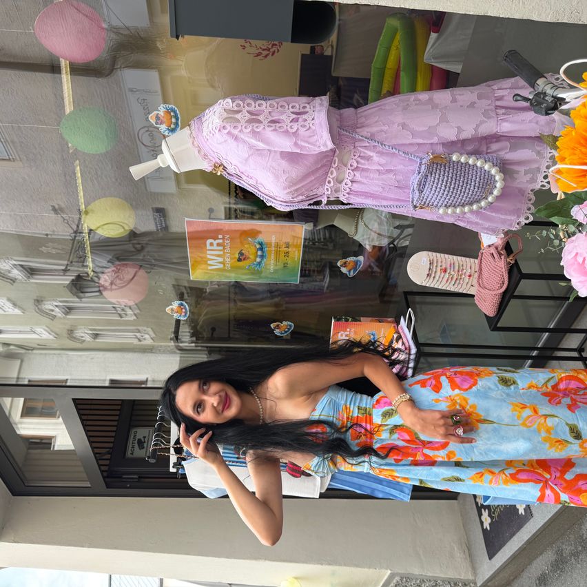 Eine Frau in einem Blumenkleid posiert vor einem Schaufenster, mit Mannequins und bunten Dekorationen im Hintergrund.