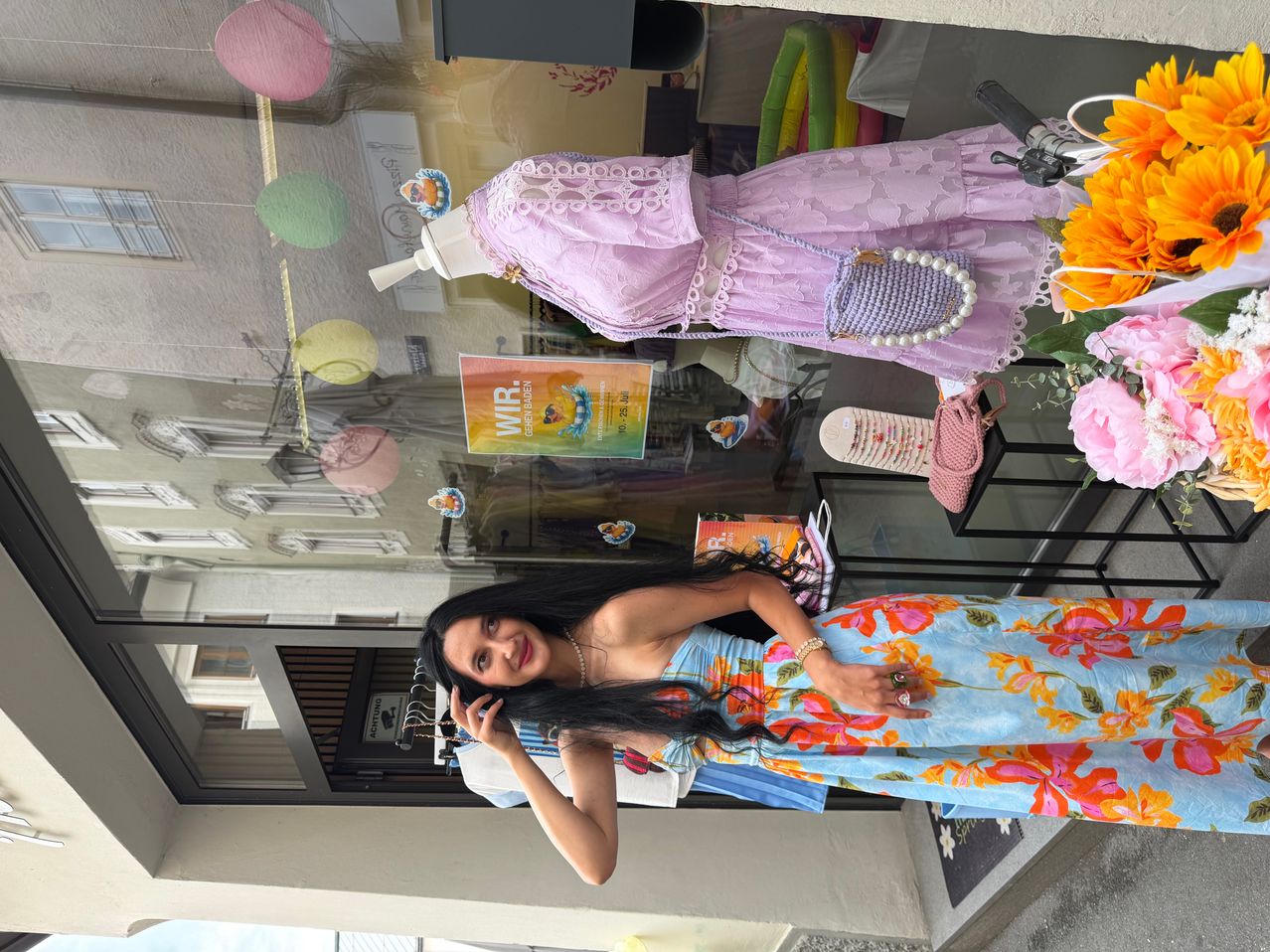 Eine Frau in einem Blumenkleid posiert vor einem Schaufenster, mit Mannequins und bunten Dekorationen im Hintergrund.